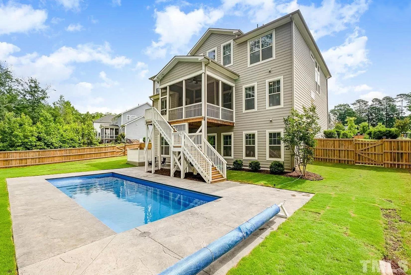 A large, green backyard with privacy fence, swimming pool, and a gray three-story home with white trim, back deck, and outdoor staircase.
